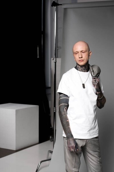 bald man with tattoos holding suit jacket on shoulder and looking away in studio