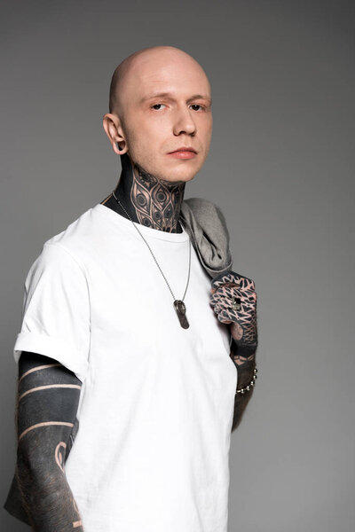 bald man with tattoos holding suit jacket on shoulder and looking at camera isolated on grey
