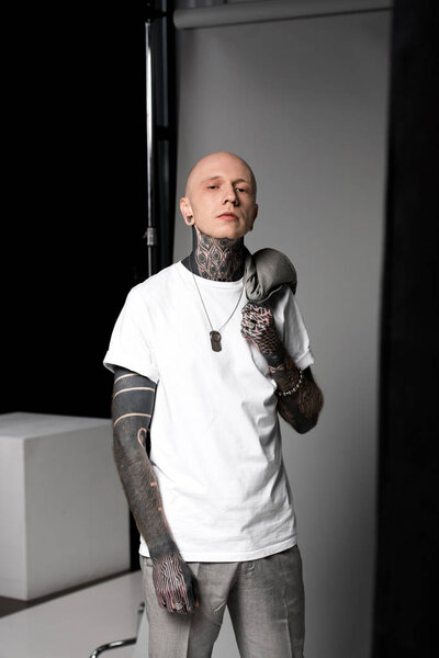 confident bald man with tattoos holding suit jacket on shoulder and looking at camera in studio