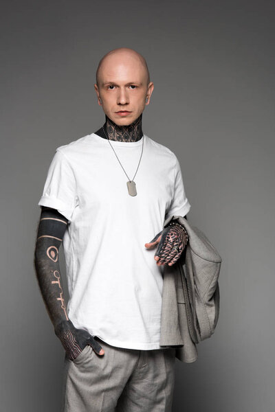 serious bald tattooed man in white t-shirt holding suit jacket and looking at camera isolated on grey 