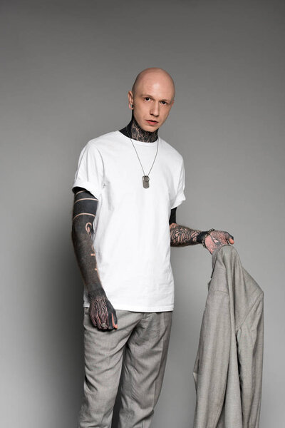 stylish bald tattooed man holding suit jacket and looking at camera on grey 
