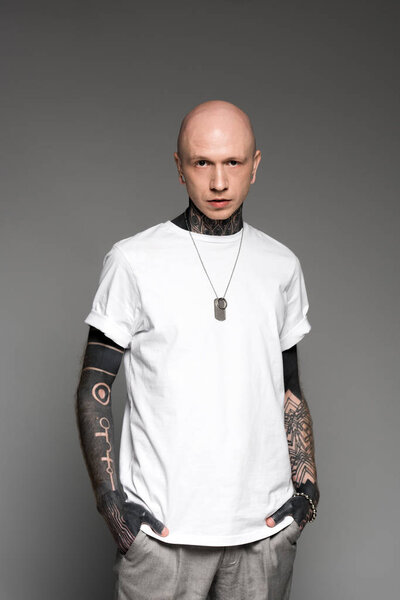 handsome bald tattooed man in white t-shirt standing with hands in pockets and looking at camera isolated on grey