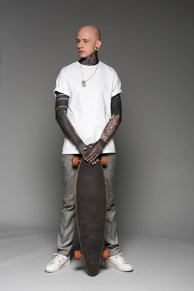 full length view of handsome bald tattooed man in white t-shirt standing with skateboard and looking away on grey