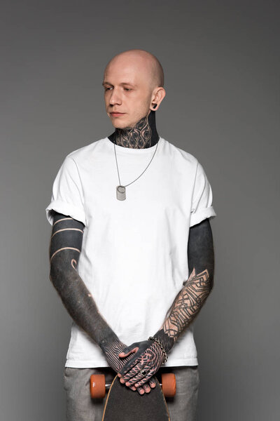 handsome bald tattooed man in white t-shirt standing with skateboard and looking away isolated on grey