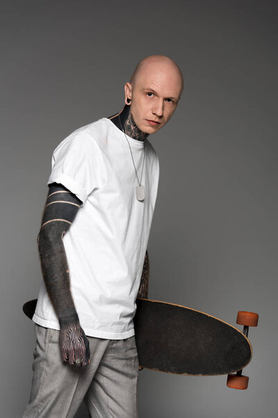 bald tattooed man in white t-shirt holding skateboard and looking at camera isolated on grey