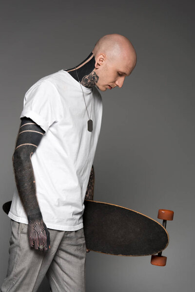 side view of bald tattooed man in white t-shirt holding skateboard and looking down isolated on grey