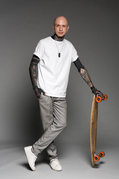 handsome tattooed man in white t-shirt standing with skateboard and looking at camera on grey