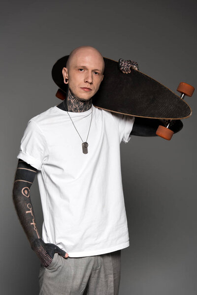 handsome tattooed man in white t-shirt holding skateboard on shoulder and looking at camera isolated on grey