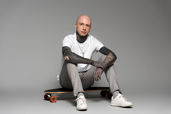 bald tattooed man sitting on skateboard and looking at camera on grey