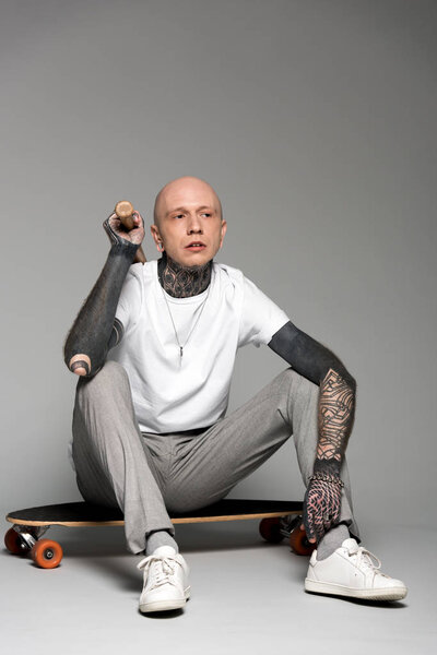 bald tattooed man sitting on skateboard and holding baseball bat on grey