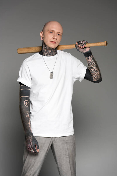 serious tattooed man in white t-shirt holding baseball bat and looking at camera isolated on grey