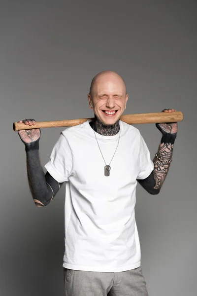 cheerful tattooed man with closed eyes standing with baseball bat on shoulders and laughing on grey