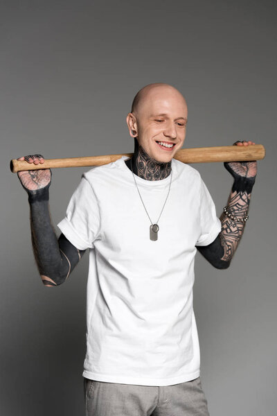 cheerful tattooed man standing with baseball bat on shoulders and laughing on grey 