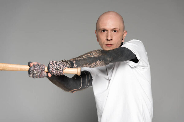 aggressive tattooed man hitting with baseball bat and looking at camera isolated on grey 