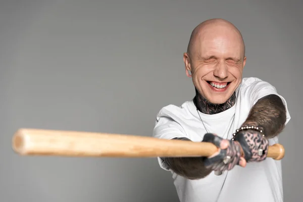 bald tattooed man with closed eyes hitting with baseball bat and laughing isolated on grey