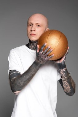 Altın basketbol topu tutarak ve gri izole kamera bakıyor beyaz t-shirt kel dövmeli adam