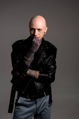 kel dövmeli adam çene üzerinde el ile deri ceket ayakta ve uzak gri izole arıyor