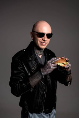 dövmeli adam deri ceket ve lezzetli burger tutarak ve gri izole kamera gülümseyen güneş gözlüğü 