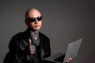 ciddi deri ceketli adamı dövmesi ve laptop ve gösteren başparmak tutarak güneş gözlüğü gri izole
