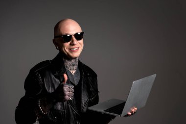 deri ceketli adamı dövmesi gülümseyen ve laptop ve gösteren başparmak tutarak güneş gözlüğü gri izole