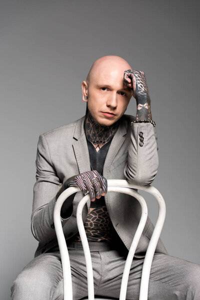 stylish tattooed man sitting on chair with hand on forehead and looking at camera isolated on grey