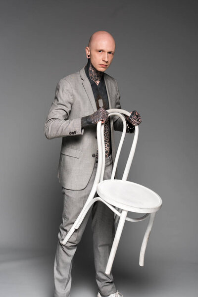 handsome serious tattooed man holding white chair and looking at camera on grey