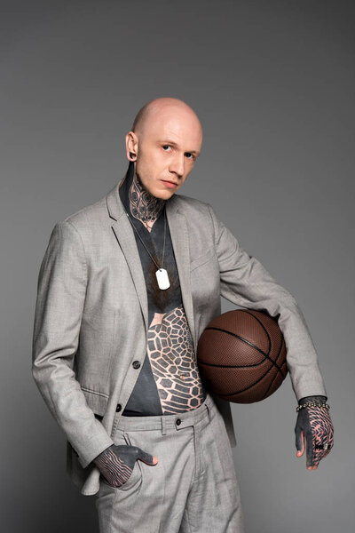 стильный лысый татуированный мужчина в костюме, держащий баскетбольный мяч и смотрящий на камеру, изолированную на сером
