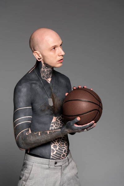 shirtless bald tattooed man holding basketball ball and looking away isolated on grey