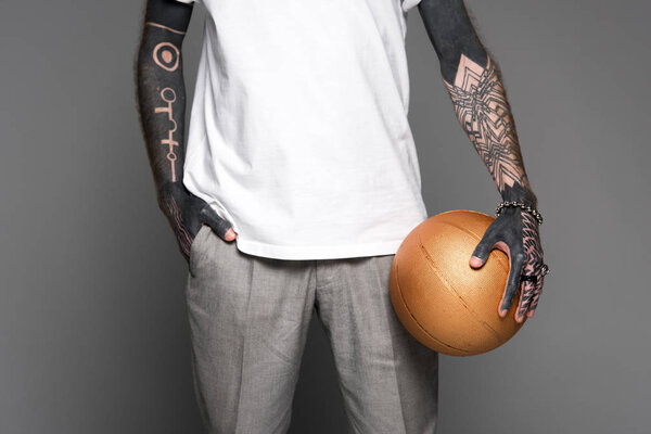 обрезанный снимок татуированного человека, стоящего с рукой в кармане и держащего золотой баскетбольный мяч, изолированный на сером
