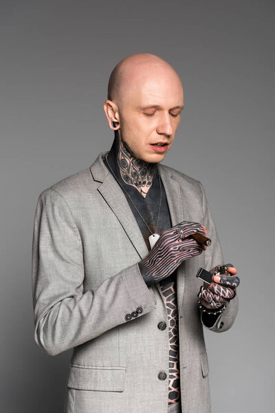 bald tattooed man in suit jacket holding cigar and lighter isolated on grey