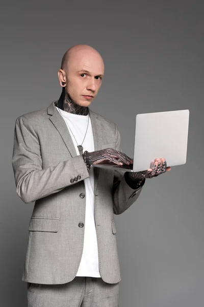 英俊的纹身男子使用笔记本电脑 看着相机孤立在灰色 — 图库照片