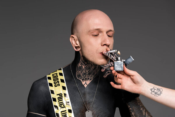partial view of someone holding lighter and tattooed man with cross line around body smoking roll-up cigarette isolated on grey