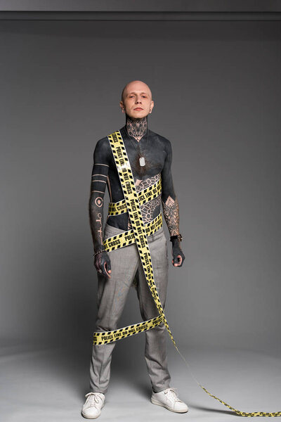 full length view of bald shirtless tattooed man with police line around body looking at camera on grey