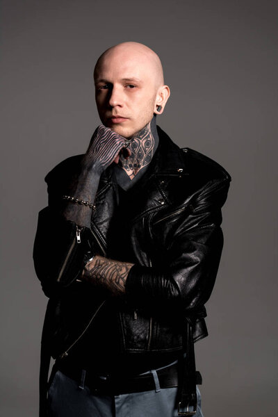 confident tattooed man in leather jacket standing with hand on chin and looking at camera isolated on grey 