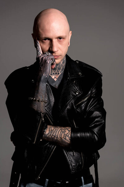 bald tattooed man in leather jacket standing with hand on face and looking at camera isolated on grey