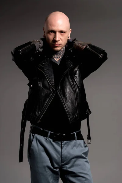 stylish tattooed man in leather jacket standing with hands behind head and looking at camera isolated on grey
