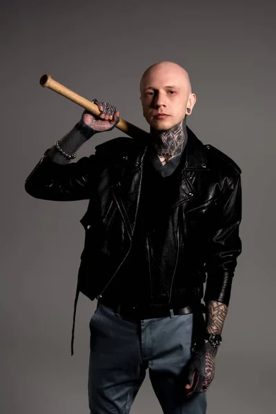 bald tattooed man in leather jacket holding baseball bat and looking at camera isolated on grey