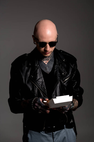 bald tattooed man in leather jacket and sunglasses holding box with tasty burger isolated on grey 