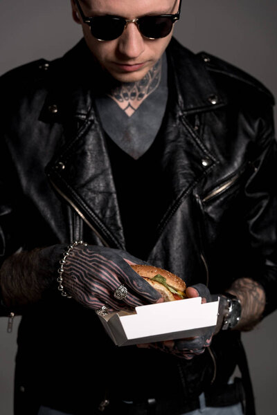 cropped shot of tattooed man in leather jacket and sunglasses holding box with tasty burger isolated on grey 