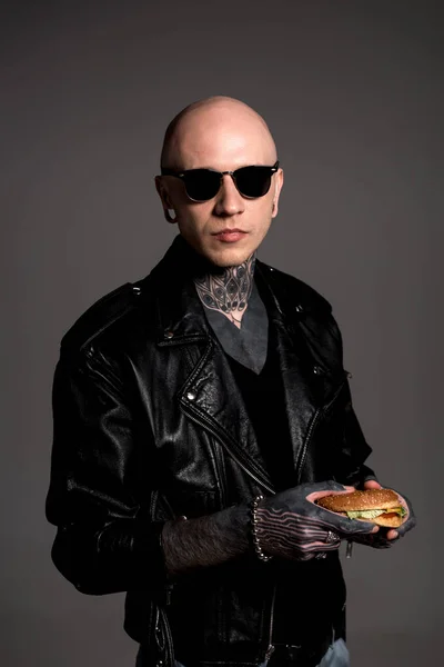纹身男子在皮夹克和太阳镜拿着美味的汉堡 看着相机孤立在灰色 — 图库照片