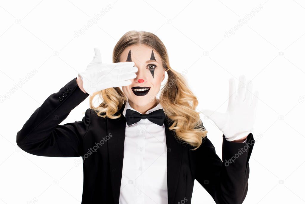 cheerful female clown standing in suit and covering eye with hand in glove isolated on white 