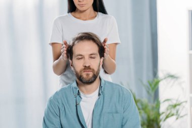 cropped shot of bearded man with closed eyes sitting and receiving reiki treatment from young female healer clipart