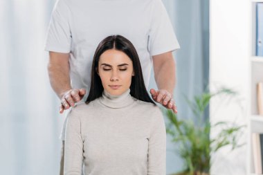 cropped shot of calm young woman with closed eyes receiving reiki healing treatment on shoulders