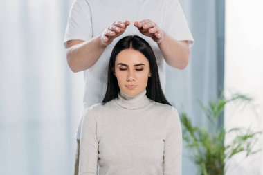 cropped shot of calm young woman with closed eyes receiving reiki healing treatment above head  clipart