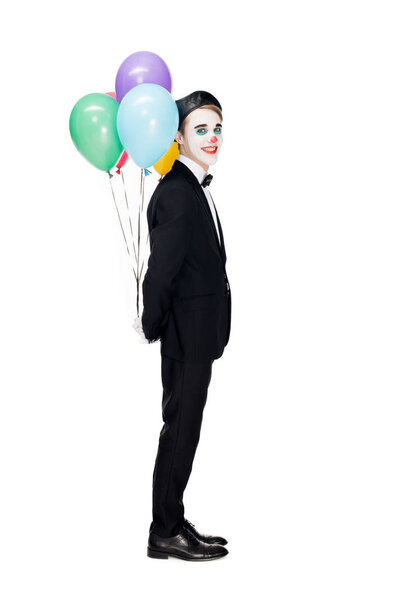 cheerful clown in suit and black beret holding helium balloons behind back isolated on white 