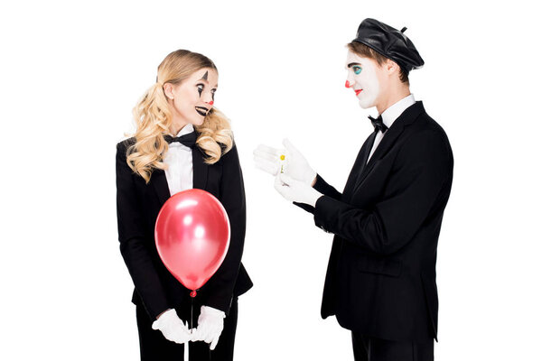 smiling female clown holding balloon and looking at man with flower isolated on white 