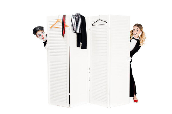smiling woman in suit looking out of dressing room door near surprised clown isolated on white 