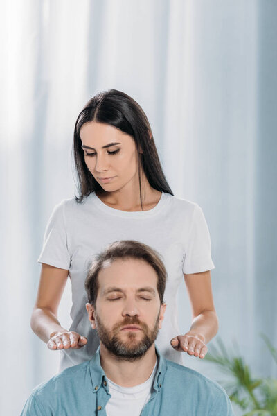 young woman doing reiki therapy session to bearded calm man with closed eyes