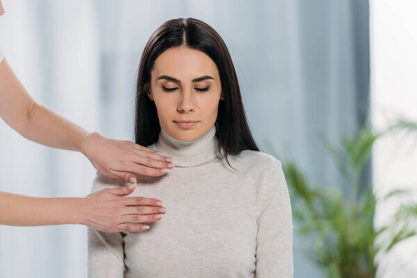 обрезанный снимок мирной молодой женщины с закрытыми глазами, сидящей и получающей лечение рейки
