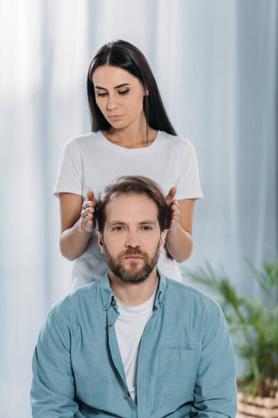 bearded man looking at camera while receiving reiki treatment from young female healer 